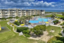 A coastal resort scene featuring a large swimming pool surrounded by sun loungers and umbrellas. Adjacent to the pool is a multi-story building with balconies overlooking the area. The landscape includes well-maintained lawns, various plants, and a playground. In the background, the ocean and a sandy beach are visible under a partly cloudy sky.