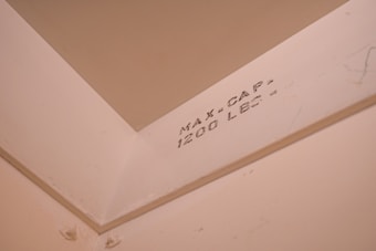 An interior corner of a room features a beige wall with a visible weight capacity indication marked 'MAX CAP - 1200 LBS -'. The surface appears smooth and painted, with lighting giving it a warm hue.