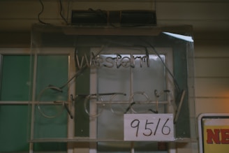 A dimly lit, old-fashioned shop sign displaying the words 'Western Grocery' is partially obscured by a white paper with numbers '95/6' written on it. The sign is mounted on a wall with a green window in the background, and there is an additional poster partially visible at the bottom right corner.