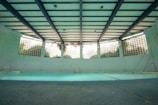 An indoor swimming pool with a turquoise floor and muted lighting. The pool is surrounded by walls with glass blocks that let in natural light. Overhead, the ceiling is structured with beams, giving the space an industrial look. There are metal ladders at the corners, and the pool area appears quiet and still.