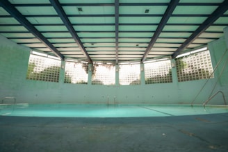 An indoor swimming pool with a turquoise floor and muted lighting. The pool is surrounded by walls with glass blocks that let in natural light. Overhead, the ceiling is structured with beams, giving the space an industrial look. There are metal ladders at the corners, and the pool area appears quiet and still.