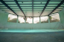 An indoor swimming pool with a turquoise floor and muted lighting. The pool is surrounded by walls with glass blocks that let in natural light. Overhead, the ceiling is structured with beams, giving the space an industrial look. There are metal ladders at the corners, and the pool area appears quiet and still.