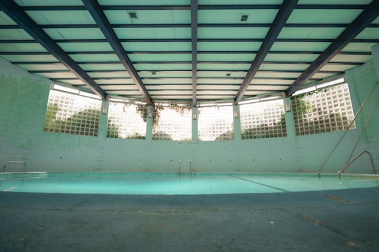 An indoor swimming pool with a turquoise floor and muted lighting. The pool is surrounded by walls with glass blocks that let in natural light. Overhead, the ceiling is structured with beams, giving the space an industrial look. There are metal ladders at the corners, and the pool area appears quiet and still.