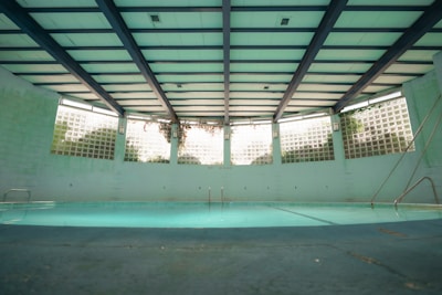 An indoor swimming pool with a turquoise floor and muted lighting. The pool is surrounded by walls with glass blocks that let in natural light. Overhead, the ceiling is structured with beams, giving the space an industrial look. There are metal ladders at the corners, and the pool area appears quiet and still.