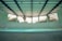 An indoor swimming pool with a turquoise floor and muted lighting. The pool is surrounded by walls with glass blocks that let in natural light. Overhead, the ceiling is structured with beams, giving the space an industrial look. There are metal ladders at the corners, and the pool area appears quiet and still.