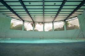 An indoor swimming pool with a turquoise floor and muted lighting. The pool is surrounded by walls with glass blocks that let in natural light. Overhead, the ceiling is structured with beams, giving the space an industrial look. There are metal ladders at the corners, and the pool area appears quiet and still.