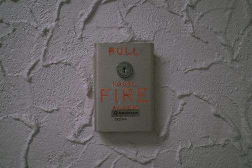 A fire alarm pull station is mounted on a textured wall. The device is metallic with bright red text indicating 'PULL' and 'LOCAL FIRE ALARM,' displaying a clear instruction for emergency use.