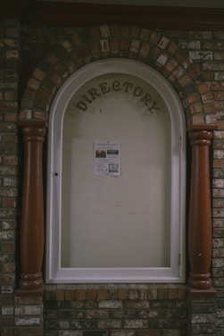 A directory display encased in an arched brick structure with a glass cover. The word 'DIRECTORY' is prominently displayed at the top, and a single sheet with information is posted inside. The surrounding brickwork features a mix of reddish and brown bricks, with two large wooden columns on either side of the glass display.