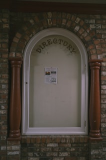 A directory display encased in an arched brick structure with a glass cover. The word 'DIRECTORY' is prominently displayed at the top, and a single sheet with information is posted inside. The surrounding brickwork features a mix of reddish and brown bricks, with two large wooden columns on either side of the glass display.