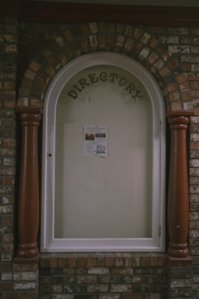 A directory display encased in an arched brick structure with a glass cover. The word 'DIRECTORY' is prominently displayed at the top, and a single sheet with information is posted inside. The surrounding brickwork features a mix of reddish and brown bricks, with two large wooden columns on either side of the glass display.