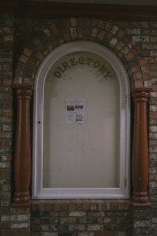 A directory display encased in an arched brick structure with a glass cover. The word 'DIRECTORY' is prominently displayed at the top, and a single sheet with information is posted inside. The surrounding brickwork features a mix of reddish and brown bricks, with two large wooden columns on either side of the glass display.