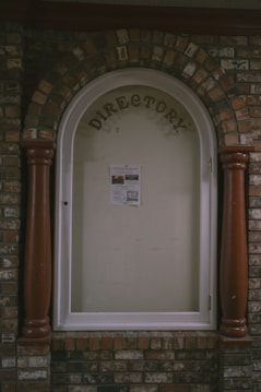 A directory display encased in an arched brick structure with a glass cover. The word 'DIRECTORY' is prominently displayed at the top, and a single sheet with information is posted inside. The surrounding brickwork features a mix of reddish and brown bricks, with two large wooden columns on either side of the glass display.