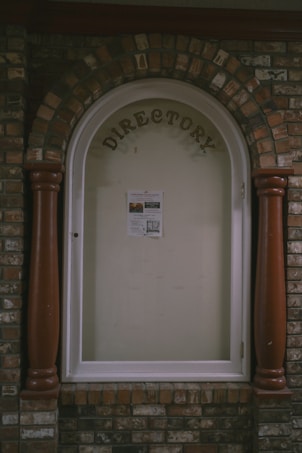 A directory display encased in an arched brick structure with a glass cover. The word 'DIRECTORY' is prominently displayed at the top, and a single sheet with information is posted inside. The surrounding brickwork features a mix of reddish and brown bricks, with two large wooden columns on either side of the glass display.