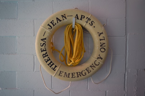 A beige life preserver ring with black text hangs on a light gray brick wall. Attached to the preserver is a coiled yellow rope. The text reads 'THERESA JEAN APTS. EMERGENCY ONLY.'