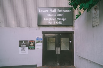 An entrance to a mall is depicted with a sign above the doors reading 'Lower Mall Entrance Fitness 2000 Lougheed Village Bar & Grill'. The area includes signs and a QR code to 'Scan to Pay' near the entrance. The surroundings are minimalistic with some greenery visible.