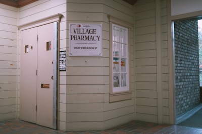 Entrance to a pharmacy with beige wooden siding. A sign reads 'Village Pharmacy' above the address '9537 Erickson Dr'. The window displays various payment logos including Visa and MasterCard, and there's a small sign indicating store hours.