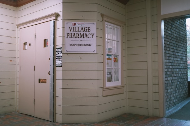 Entrance to a pharmacy with beige wooden siding. A sign reads 'Village Pharmacy' above the address '9537 Erickson Dr'. The window displays various payment logos including Visa and MasterCard, and there's a small sign indicating store hours.