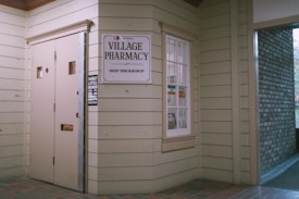 Entrance to a pharmacy with beige wooden siding. A sign reads 'Village Pharmacy' above the address '9537 Erickson Dr'. The window displays various payment logos including Visa and MasterCard, and there's a small sign indicating store hours.
