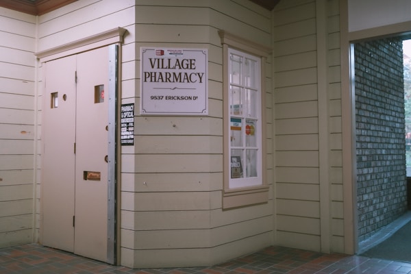 Entrance to a pharmacy with beige wooden siding. A sign reads 'Village Pharmacy' above the address '9537 Erickson Dr'. The window displays various payment logos including Visa and MasterCard, and there's a small sign indicating store hours.