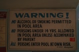 A sign displaying pool area rules, prohibiting alcohol and smoking, requiring under 14s to be accompanied by a parent, and advising all persons enter at their own risk. The sign is predominantly white and blue with bold lettering.