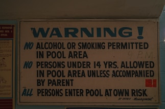 A sign displaying pool area rules, prohibiting alcohol and smoking, requiring under 14s to be accompanied by a parent, and advising all persons enter at their own risk. The sign is predominantly white and blue with bold lettering.