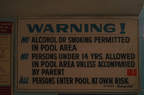 A sign displaying pool area rules, prohibiting alcohol and smoking, requiring under 14s to be accompanied by a parent, and advising all persons enter at their own risk. The sign is predominantly white and blue with bold lettering.