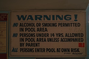 A sign displaying pool area rules, prohibiting alcohol and smoking, requiring under 14s to be accompanied by a parent, and advising all persons enter at their own risk. The sign is predominantly white and blue with bold lettering.