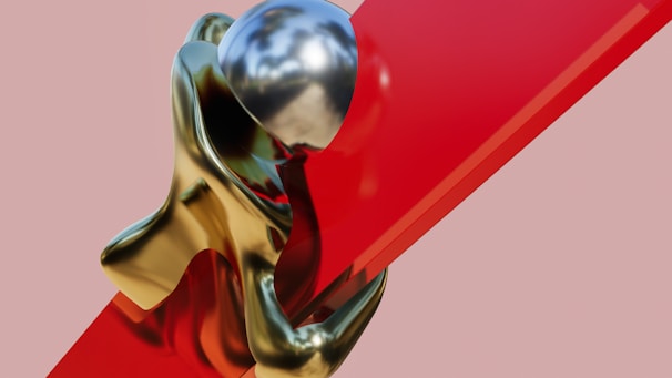 A surrealist sculpture featuring a metallic humanoid figure embracing a large red glass-like slab. The figure has a smooth shiny texture, reflecting light and the surrounding environment. The contrast between the metallic gold of the figure and the vibrant red of the slab creates a striking visual effect.
