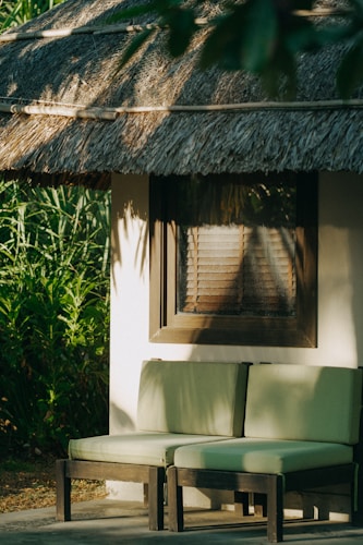 A shaded outdoor seating area with two green cushioned chairs placed against a wall of a building. The building has a thatched roof and a wooden framed window with horizontal blinds. Surrounding foliage adds a touch of greenery to the setting, which is bathed in warm sunlight.