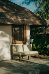 A serene outdoor setting with a thatched-roof building rendered in soft sunlight. Two cushioned chairs rest against a wall with a wooden-framed window. Surrounding greenery provides a lush backdrop, while dappled shadows play across the ground and walls.