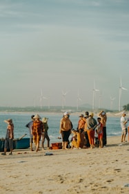 A group of people gathered on a sandy beach near the ocean. They appear to be engaged in various activities, possibly relating to fishing, as there is a boat and some crates nearby. Some are wearing traditional conical hats, and the background features wind turbines along the coastline.