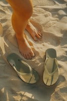 A pair of human feet, one stepping forward, are seen standing on sandy ground. Beside the feet, there is a pair of green flip-flops placed on the sand. The texture of the sand and the shadows cast by the legs and flip-flops are clearly visible.