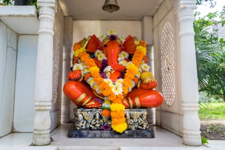 A vibrant orange statue of the Hindu deity Ganesha adorned with an intricate garland made of orange, yellow, white, and purple flowers. The statue sits on an ornate base within a small open marble shrine surrounded by greenery.