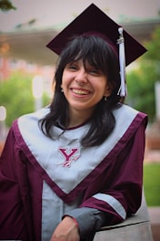 A person is wearing a maroon graduation cap and gown with a gray stole featuring a Y logo, smiling broadly. The background is blurred, showing greenery and a building.
