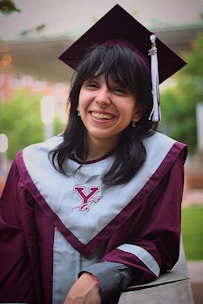 A person is wearing a maroon graduation cap and gown with a gray stole featuring a Y logo, smiling broadly. The background is blurred, showing greenery and a building.