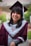 A person is wearing a maroon graduation cap and gown with a gray stole featuring a Y logo, smiling broadly. The background is blurred, showing greenery and a building.