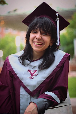 A person is wearing a maroon graduation cap and gown with a gray stole featuring a Y logo, smiling broadly. The background is blurred, showing greenery and a building.