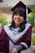 A person is wearing a maroon graduation cap and gown with a gray stole featuring a Y logo, smiling broadly. The background is blurred, showing greenery and a building.