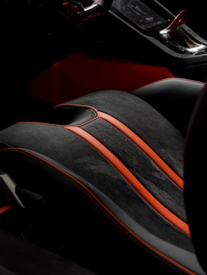 A detailed view of a car's interior, showcasing a high-end seat with a black and red color scheme. The seat features sleek black leather with vibrant red accents and stitching, highlighting a luxurious and sporty design. Part of the console is visible, emphasizing modern and sophisticated aesthetics.