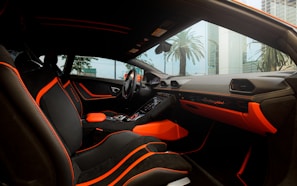 The interior of a luxury car featuring black seating with bright orange accents and stitching. The dashboard displays the brand logo in elegant script, also highlighted with orange elements. The design is sleek and modern, with large windows capturing an external view of palm trees and high-rise buildings.