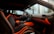 The interior of a luxury car featuring black seating with bright orange accents and stitching. The dashboard displays the brand logo in elegant script, also highlighted with orange elements. The design is sleek and modern, with large windows capturing an external view of palm trees and high-rise buildings.