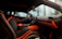 The interior of a luxury car featuring black seating with bright orange accents and stitching. The dashboard displays the brand logo in elegant script, also highlighted with orange elements. The design is sleek and modern, with large windows capturing an external view of palm trees and high-rise buildings.
