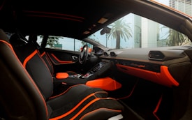 The interior of a luxury car featuring black seating with bright orange accents and stitching. The dashboard displays the brand logo in elegant script, also highlighted with orange elements. The design is sleek and modern, with large windows capturing an external view of palm trees and high-rise buildings.