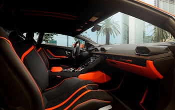 The interior of a luxury car featuring black seating with bright orange accents and stitching. The dashboard displays the brand logo in elegant script, also highlighted with orange elements. The design is sleek and modern, with large windows capturing an external view of palm trees and high-rise buildings.