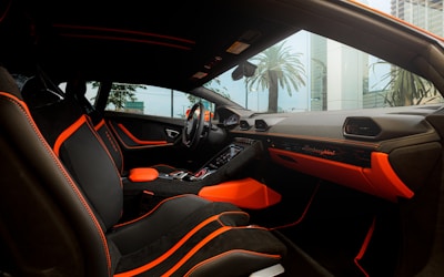 The interior of a luxury car featuring black seating with bright orange accents and stitching. The dashboard displays the brand logo in elegant script, also highlighted with orange elements. The design is sleek and modern, with large windows capturing an external view of palm trees and high-rise buildings.