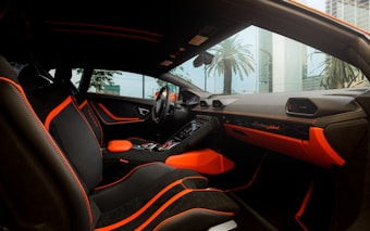 The interior of a luxury car featuring black seating with bright orange accents and stitching. The dashboard displays the brand logo in elegant script, also highlighted with orange elements. The design is sleek and modern, with large windows capturing an external view of palm trees and high-rise buildings.