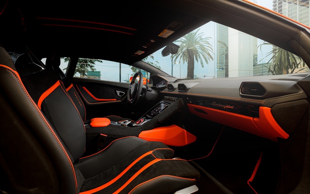 The interior of a luxury car featuring black seating with bright orange accents and stitching. The dashboard displays the brand logo in elegant script, also highlighted with orange elements. The design is sleek and modern, with large windows capturing an external view of palm trees and high-rise buildings.
