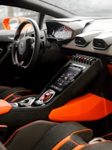 A luxurious car interior featuring a modern dashboard with digital displays, sleek steering wheel, and vibrant orange and black upholstery. The center console includes advanced electronics and controls, highlighting a sporty and high-tech design.