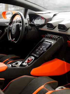 A luxurious car interior featuring a modern dashboard with digital displays, sleek steering wheel, and vibrant orange and black upholstery. The center console includes advanced electronics and controls, highlighting a sporty and high-tech design.