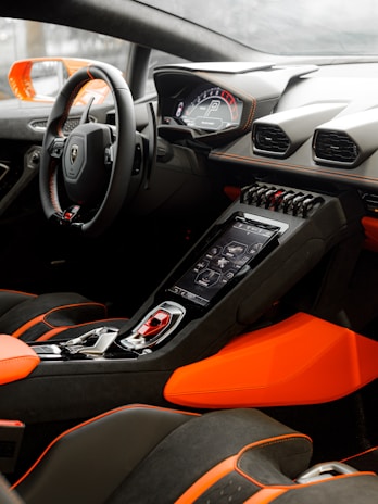 A luxurious car interior featuring a modern dashboard with digital displays, sleek steering wheel, and vibrant orange and black upholstery. The center console includes advanced electronics and controls, highlighting a sporty and high-tech design.